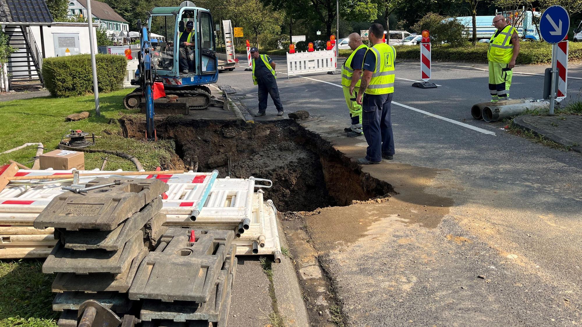 Um die Baugrube mit Bagger stehen die Arbeiter mit gelber Weste und begutachten die Leitungen im Untergrund.