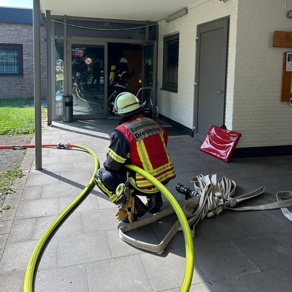 Ein Feuerwehrmann hält einen Wasserschlauch an einem Gebäude, in dem es gebrannt hat. Vor ihm gehen Feuerwehrleute durch ein Gebäude.