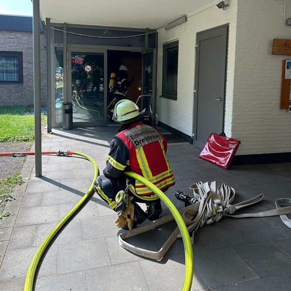 Ein Feuerwehrmann hält einen Wasserschlauch an einem Gebäude, in dem es gebrannt hat. Vor ihm gehen Feuerwehrleute durch ein Gebäude.