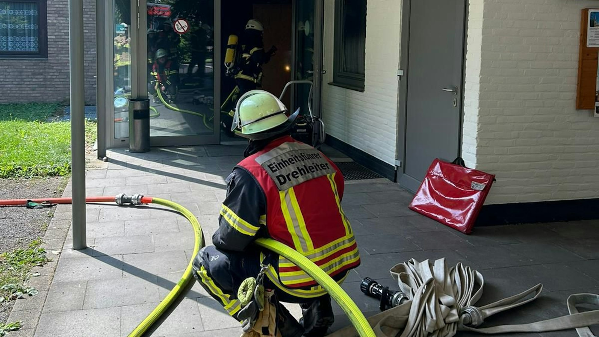 Ein Feuerwehrmann hält einen Wasserschlauch an einem Gebäude, in dem es gebrannt hat. Vor ihm gehen Feuerwehrleute durch ein Gebäude.