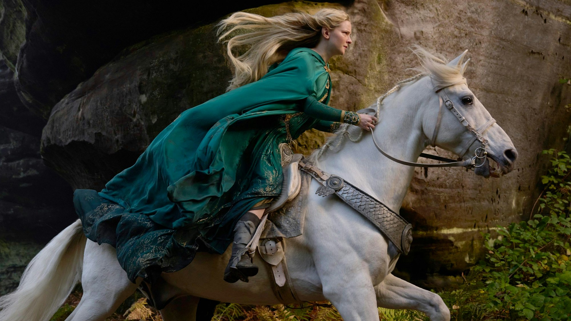 Die walisische Schauspielerin Morfydd Clark reitet in ihrer Rolle als Elbenkriegerin Galadriel in der Amazon-Prime-Video-Serie „Der Herr der Ringe: Die Ringe der Macht“ einen Schimmel. Ihre blonden Haare und ihre grünen Gewänder wehen im Wind.