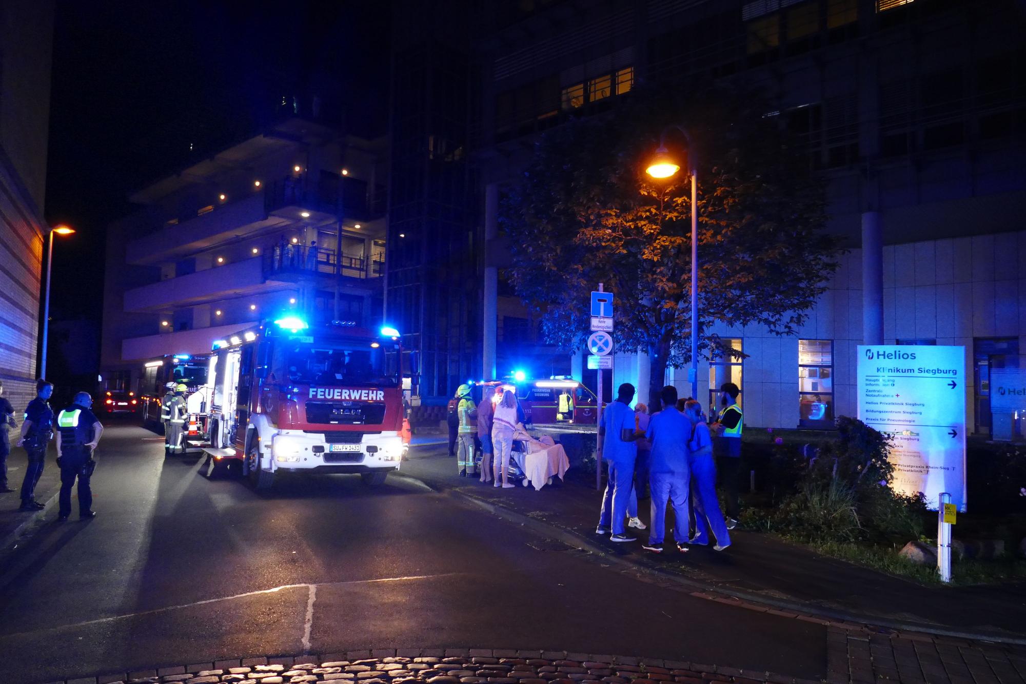 Ein Feuerwehrauto steht auf einer Straße, daneben Personen in blauen Klamotten.