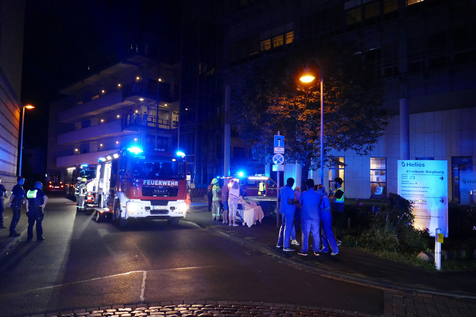 Ein Feuerwehrauto steht auf einer Straße, daneben Personen in blauen Klamotten.