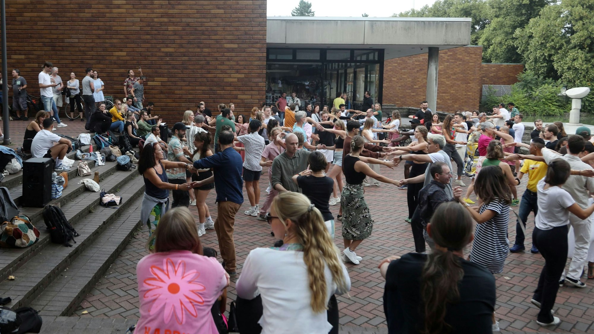 Auf der Terrasse vor dem Museum für Ostasiatische Kunst sind zahlreiche Menschen zusammengekommen, um Salsa zu tanzen. Schaulustige bevölkern sitzend und stehend die Treppen, die zur Terrasse hinunterführen.