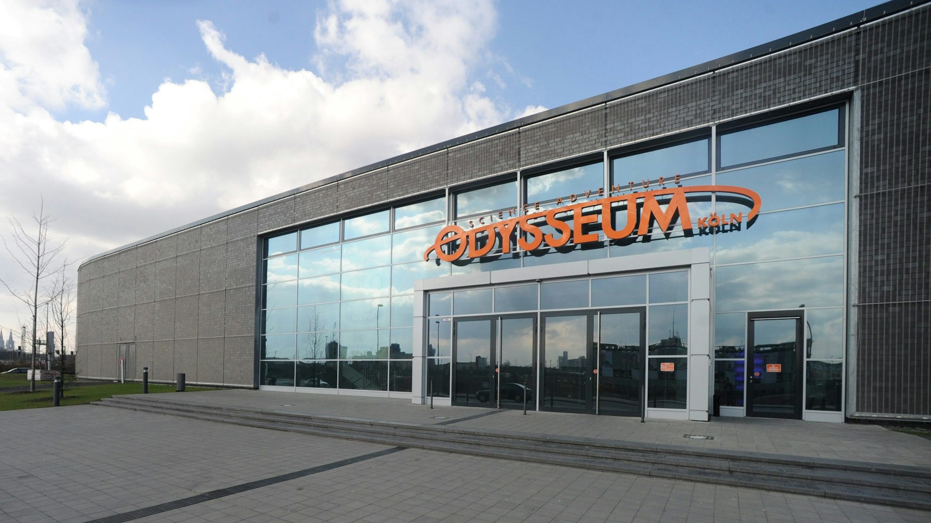 Das Foto zeigt den Eingangsbereich des Odysseum. Vom verglasten Eingangsbereich hebt sich die schmucklose graue Fassade des flachen, langgestreckten Bauwerks ab. Über dem Eingang prangt in orangefarbener Schrift der Name „Odysseum Köln“.