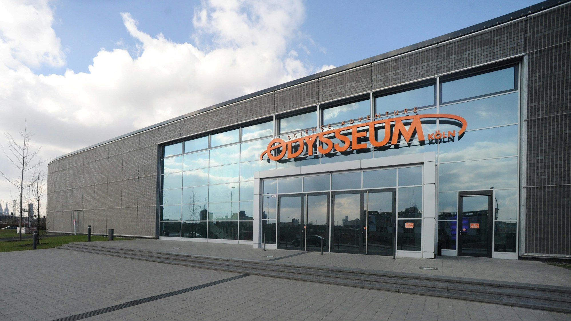Das Foto zeigt den Eingangsbereich des Odysseum. Vom verglasten Eingangsbereich hebt sich die schmucklose graue Fassade des flachen, langgestreckten Bauwerks ab. Über dem Eingang prangt in orangefarbener Schrift der Name „Odysseum Köln“.