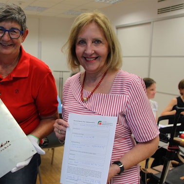 Zwei Frauen stehen in einem Raum. Eine hält eine Schablone in den Händen, die andere eine Urkunde.