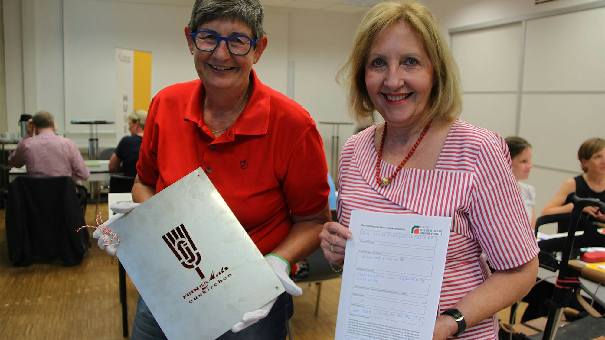 Zwei Frauen stehen in einem Raum. Eine hält eine Schablone in den Händen, die andere eine Urkunde.