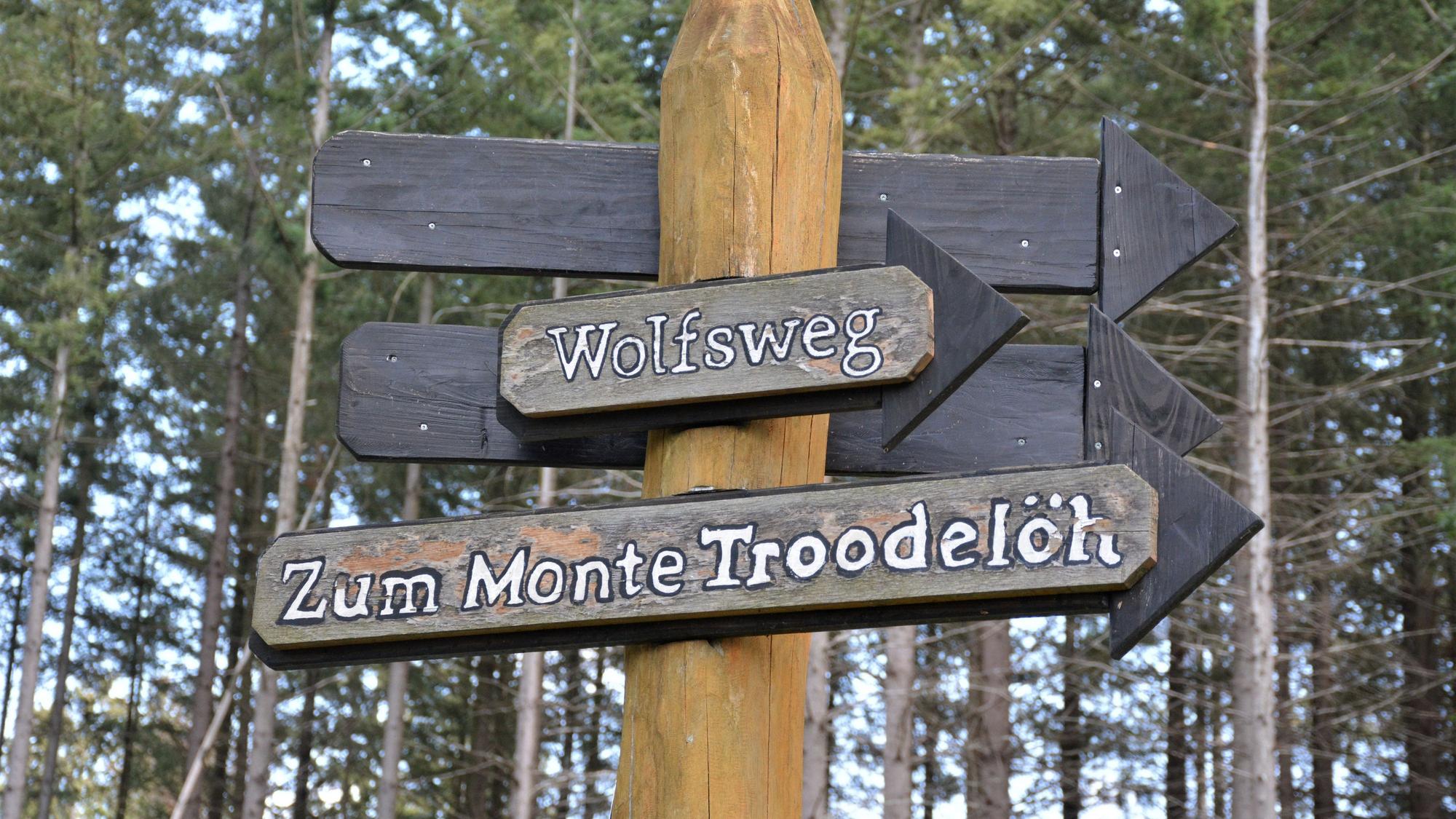 Zu sehen sind zwei Schilder in einem Wald: „Wolfsweg“ und „Zum Monte Troodelöh“.