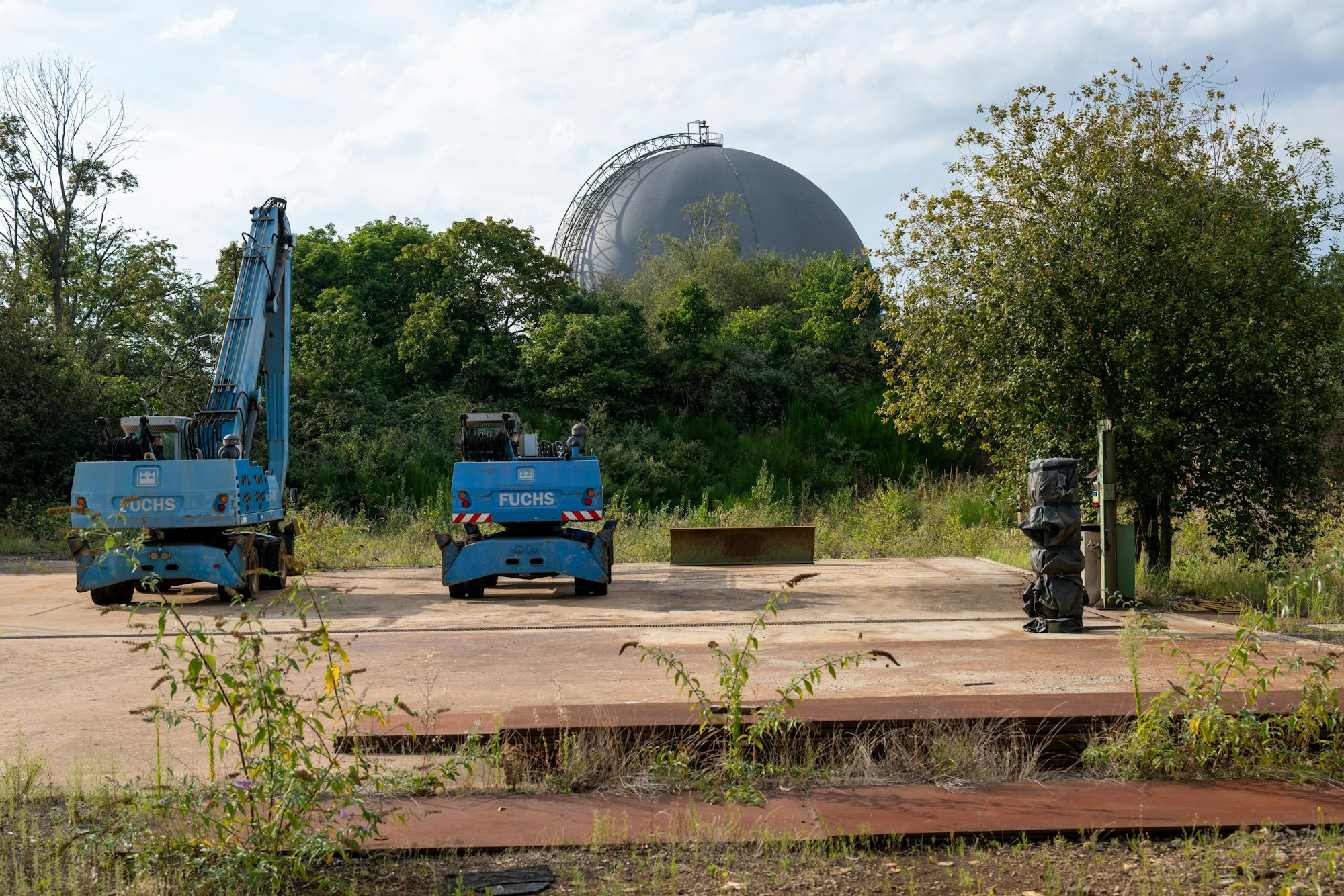 Auf dem Max-Becker-Areal finden sich noch Reste des ehemaligen Schrottplatzes: Bagger und Metall im Vordergrund, dahinter eine zugewucherte große Gaskugel.