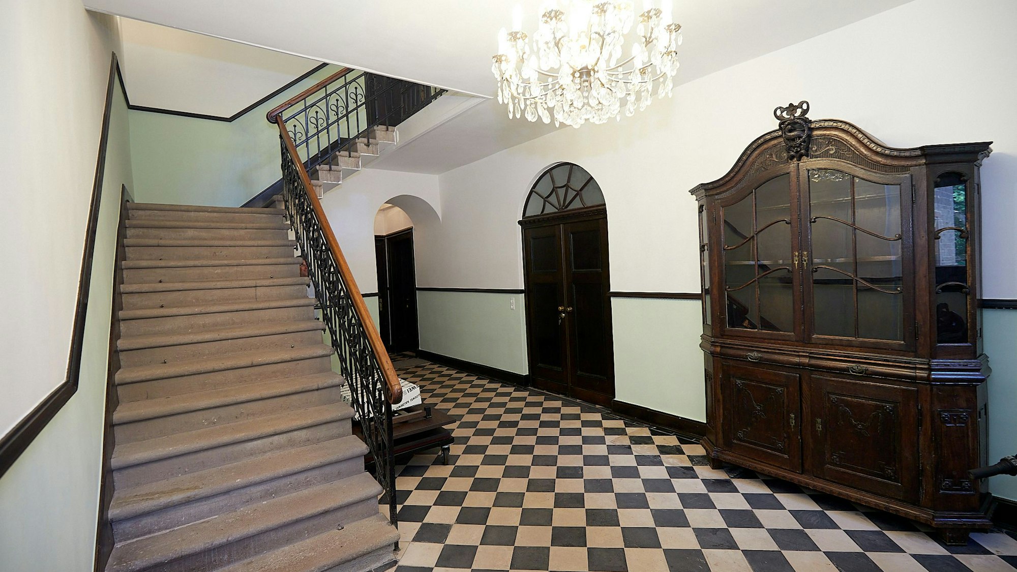 Vom Flur aus führt eine Treppe ins obere Geschoss. An einer Wand steht ein aufwendig gestalteter Schrank, an der Decke hängt ein Kronleuchter.