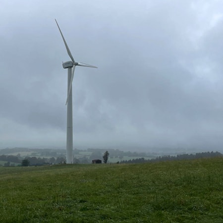 Ein einzelnes Windrad steht auf einer grünen Wiese vor wolkenverhangenem Himmel, im Hintergrund sind grüne Hügel und Wälder zu sehen.