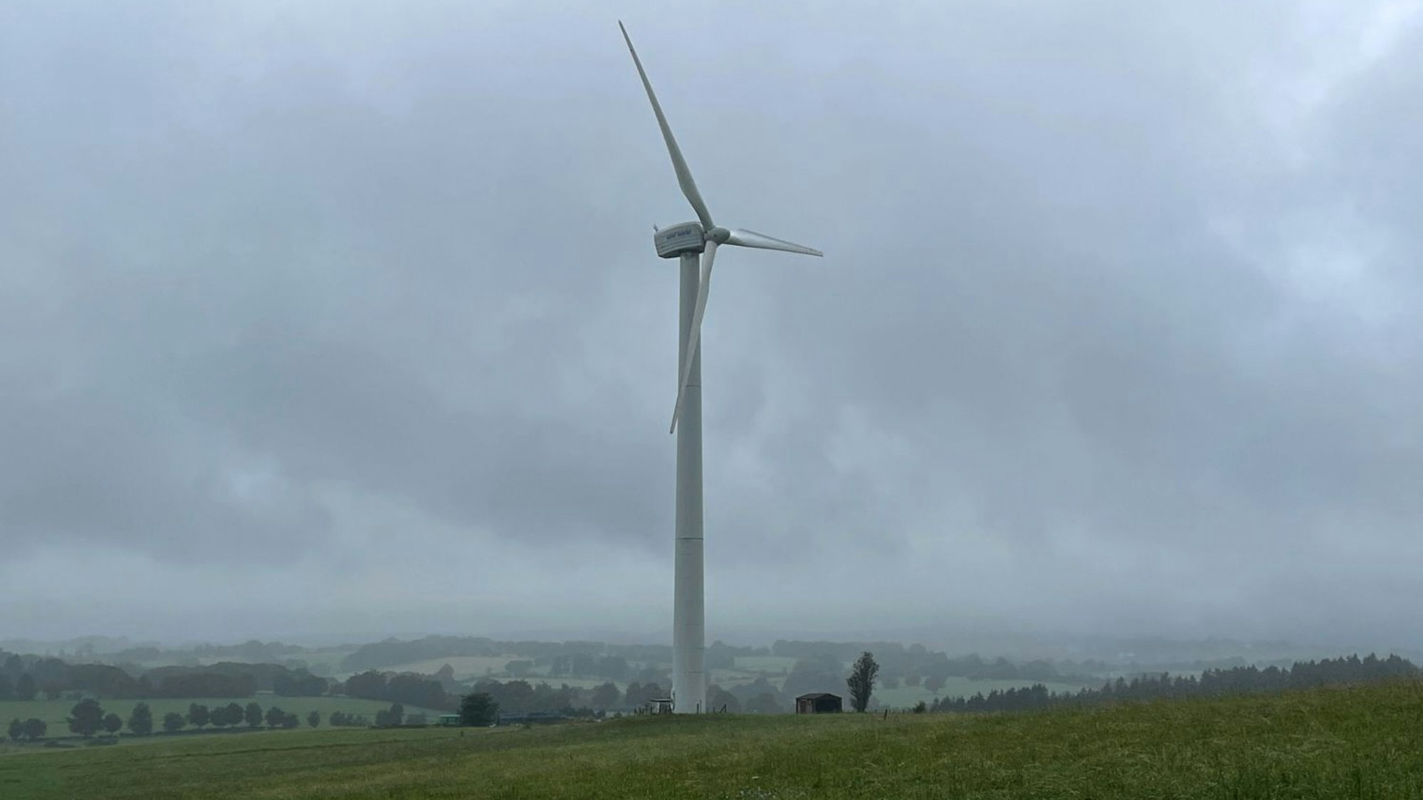 Ein einzelnes Windrad steht auf einer grünen Wiese vor wolkenverhangenem Himmel, im Hintergrund sind grüne Hügel und Wälder zu sehen.