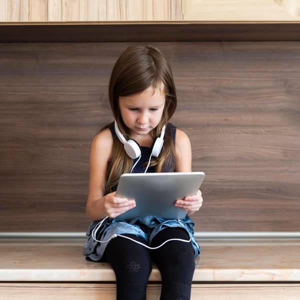Ein kleines Mädchen sitzt auf einer Bank und hält ein Tablet in der Hand. Sie trägt Kopfhörer.