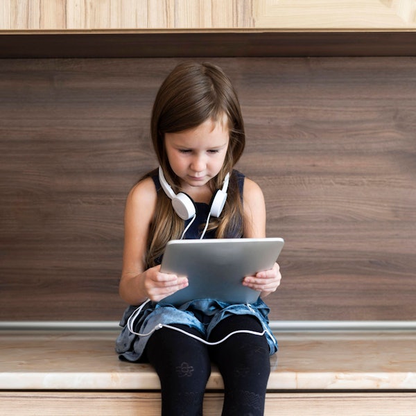 Ein kleines Mädchen sitzt auf einer Bank und hält ein Tablet in der Hand. Sie trägt Kopfhörer.