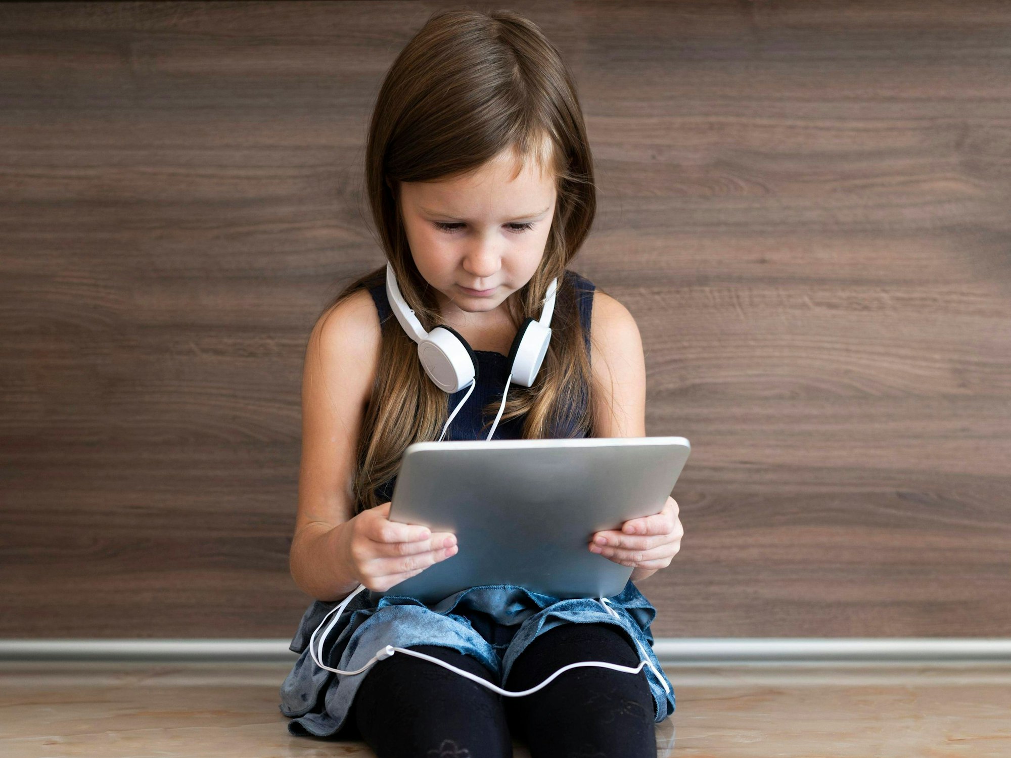Ein kleines Mädchen sitzt auf einer Bank und hält ein Tablet in der Hand. Sie trägt Kopfhörer.