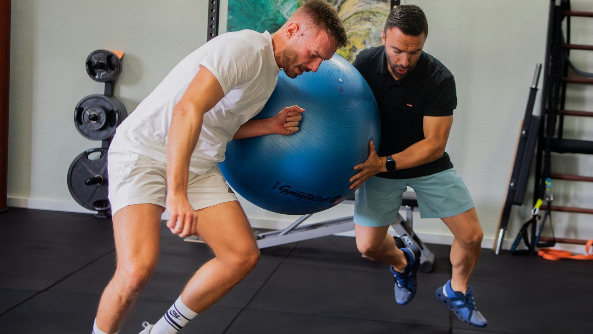 Das Bild zeigt Robin Ritz, wie er mit der linken Schulter gegen einen blauen Gummiball drückt. Der Ball wird von seinem Trainer gehalten.