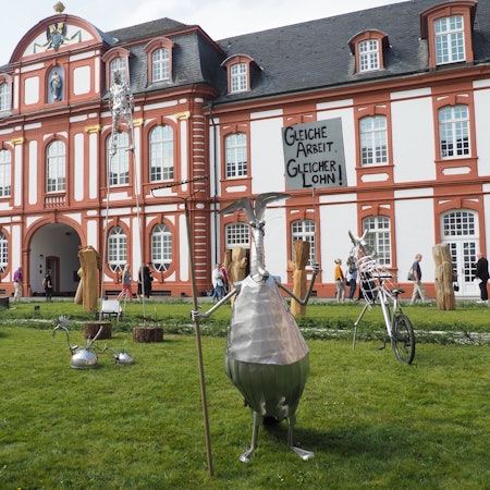 Vor der Abtei in Pulheim-Brauweiler stehen verschiedene kleine Skulpturen aus Blech. Vorne im Bild etwa ein stehender Ameisenbär, der eine Harke hält wie einen Speer.