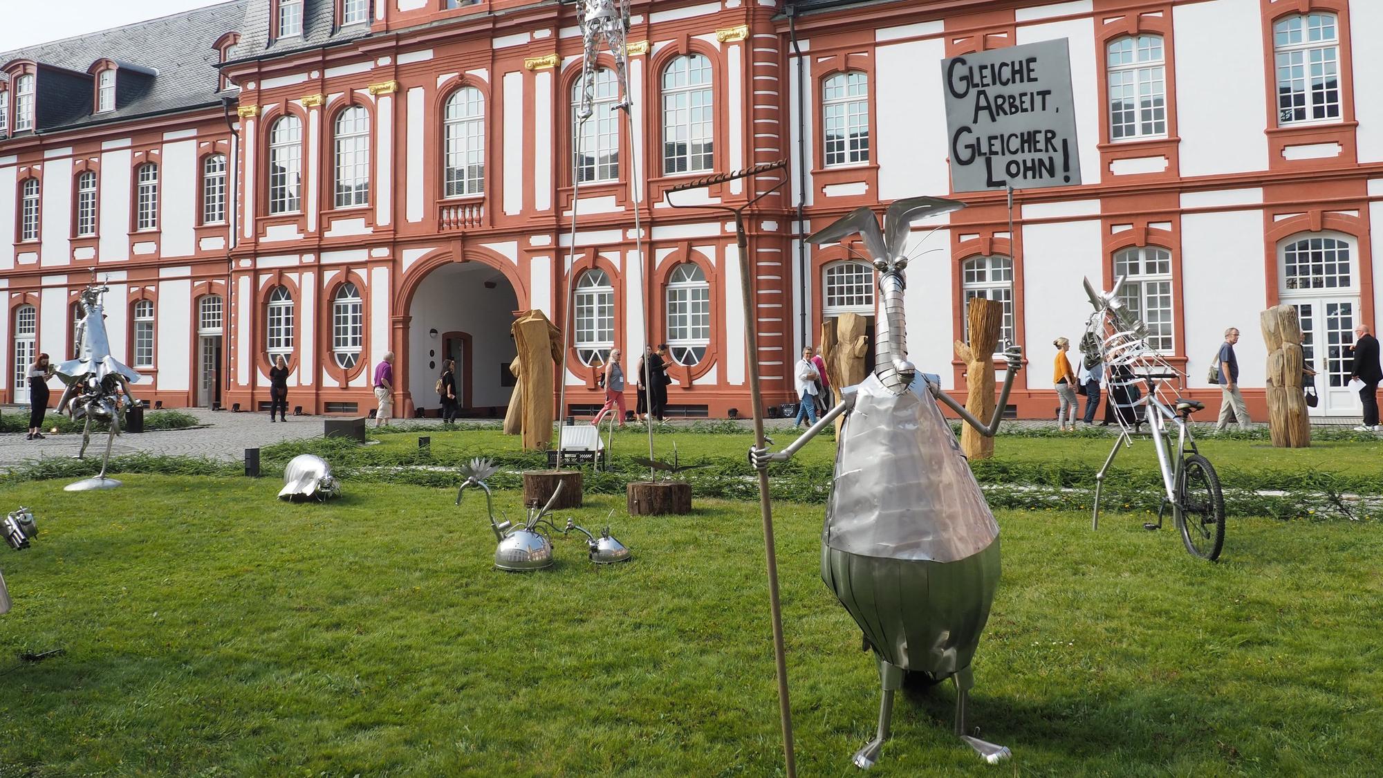 Vor der Abtei in Pulheim-Brauweiler stehen verschiedene kleine Skulpturen aus Blech. Vorne im Bild etwa ein stehender Ameisenbär, der eine Harke hält wie einen Speer.