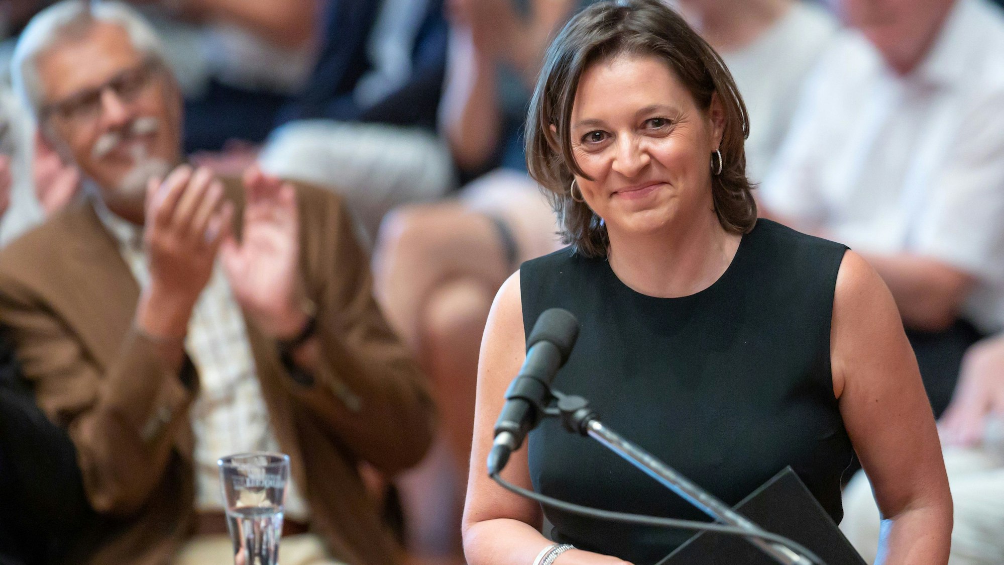 Husch Josten steht an einem Rednerpult, sie trägt ein dunkelblaues, ärmelloses Kleid.