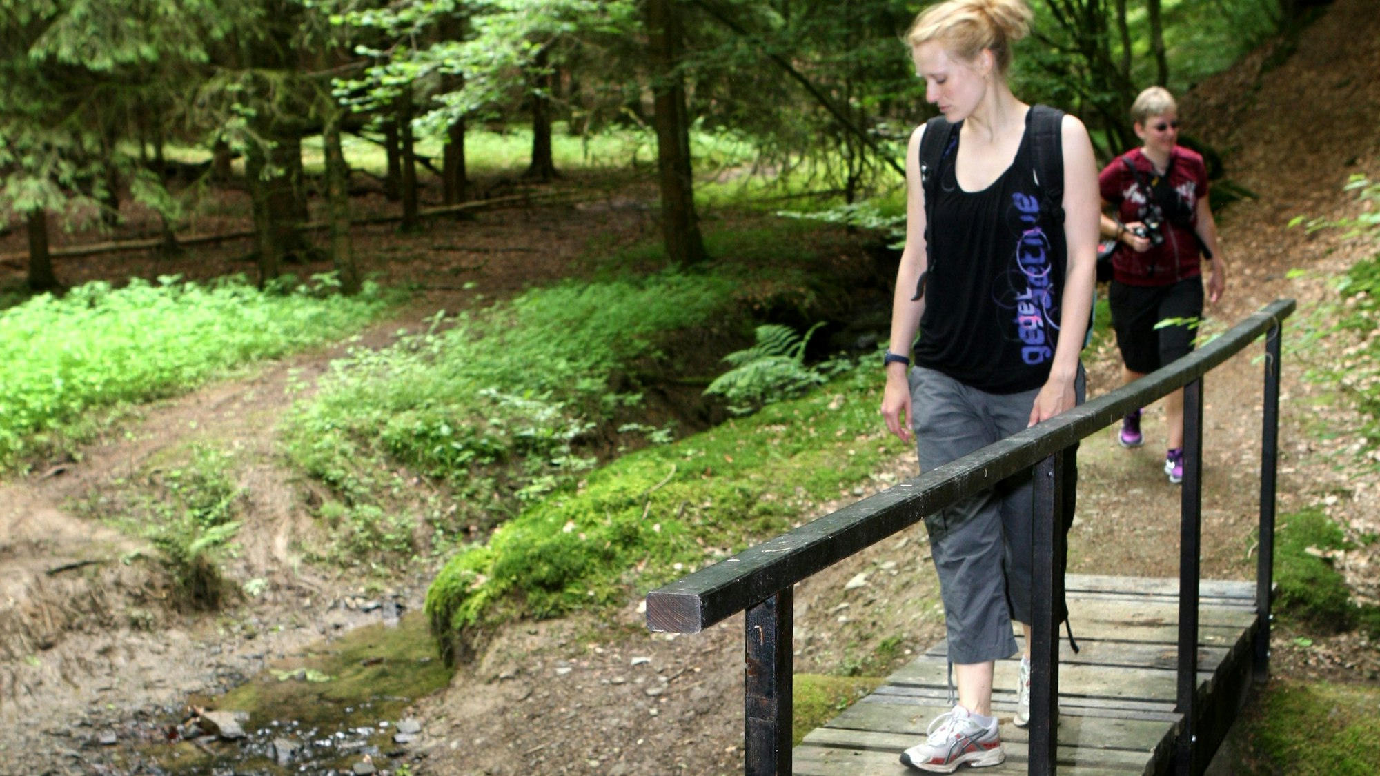 Eine Frau geht auf einem Steg über einen Bach in einem waldreichen Tal bei Neunkirchen-Seelscheid.