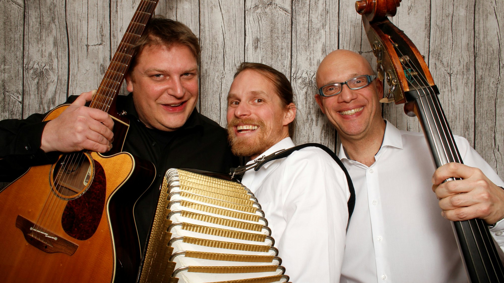 drei Musiker mit Gitarre, Akkordeon und Bass halten ihre Instrumente in die Kamera