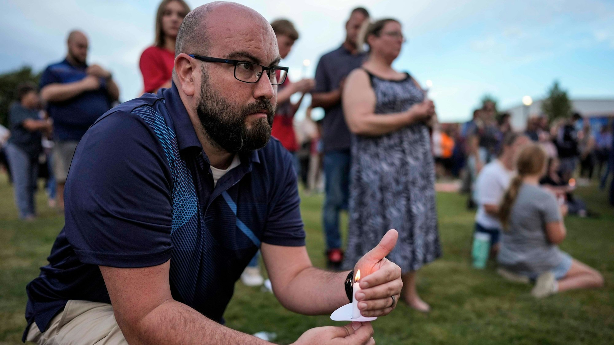 Ein Mann mit Glatze und Brille hält eine Kerze bei einer Mahnwache und kniet auf einer Wiese. Hinter ihm stehen und sitzen viele andere Teilnehmende der Mahnwache.