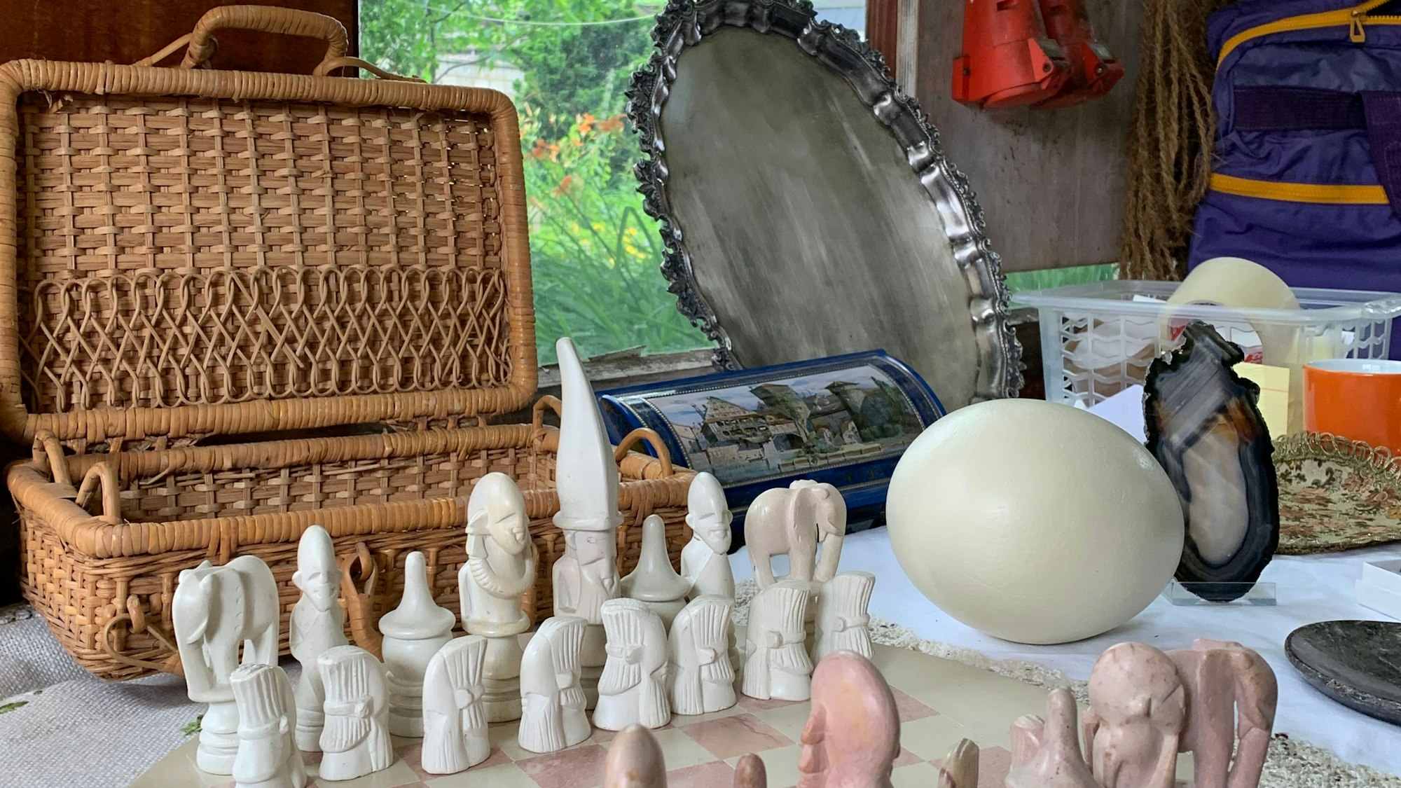 Das Bild zeigt einen Picknickkorb im Hintergrund, im Vordergrund ein Schachset. Daneben liegt ein Straußenei und weitere Gegenstände von einem Trödelmarkt.