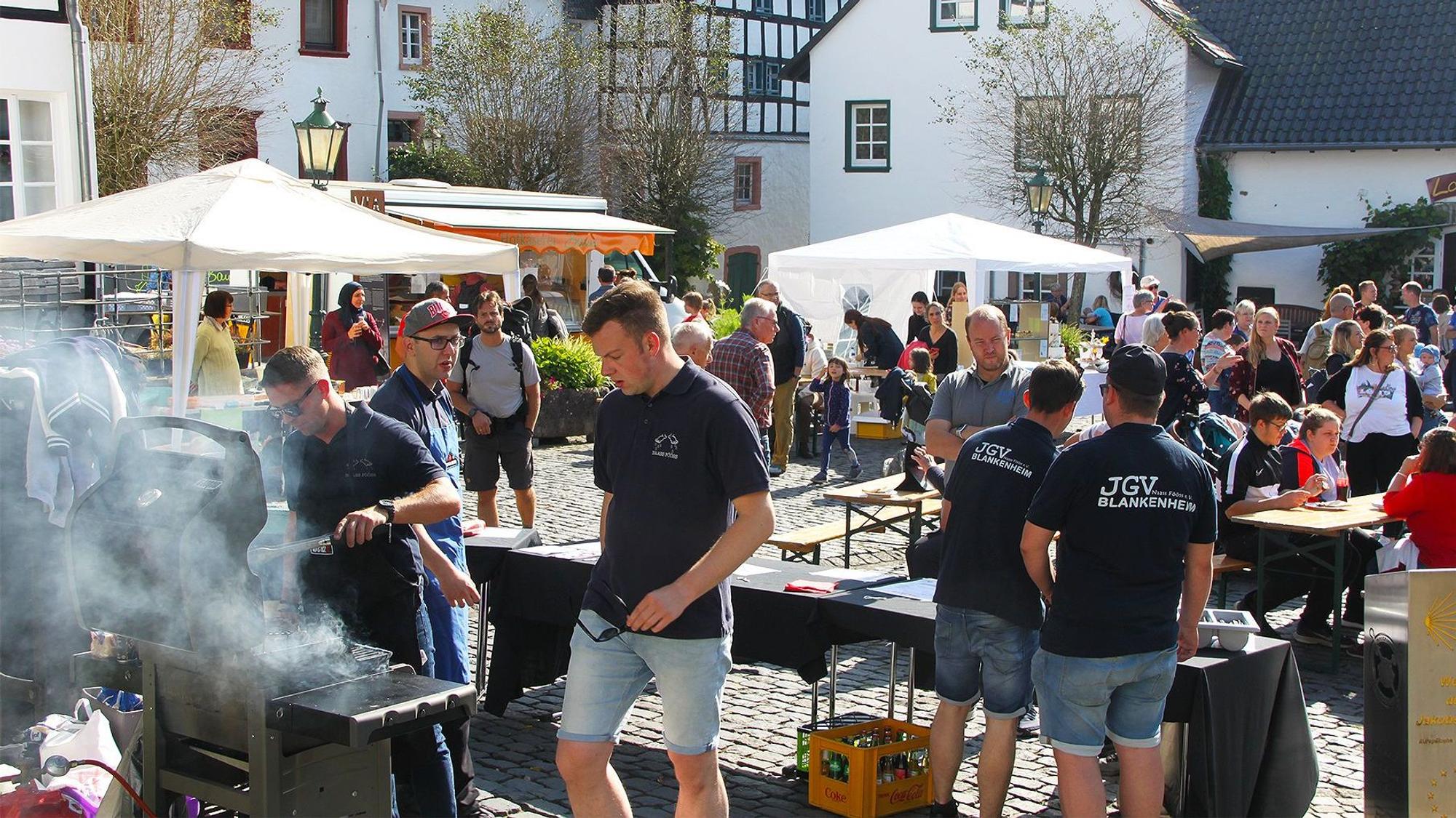 Einige junge Männer stehen in Blankenheim an einem Grill. Dahinter sind auf dem Platz zahlreiche Besucher zu erkennen.