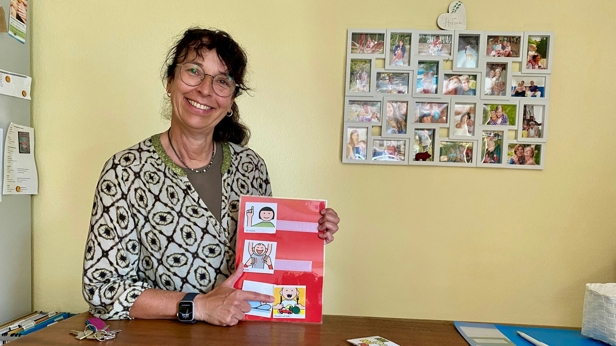 Eine Frau mit Brille zeigt grinsend auf ein Zeichentrick-Bild. Im Hintergrund hängen Fotos in einer Collage an einer Wand.