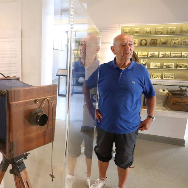 Ein Mann in blauem Poloshirt neben einer historischen Plattenkamera, die in einer Vitrine steht.