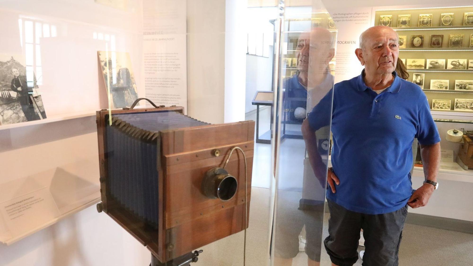 Ein Mann in blauem Poloshirt neben einer historischen Plattenkamera, die in einer Vitrine steht.