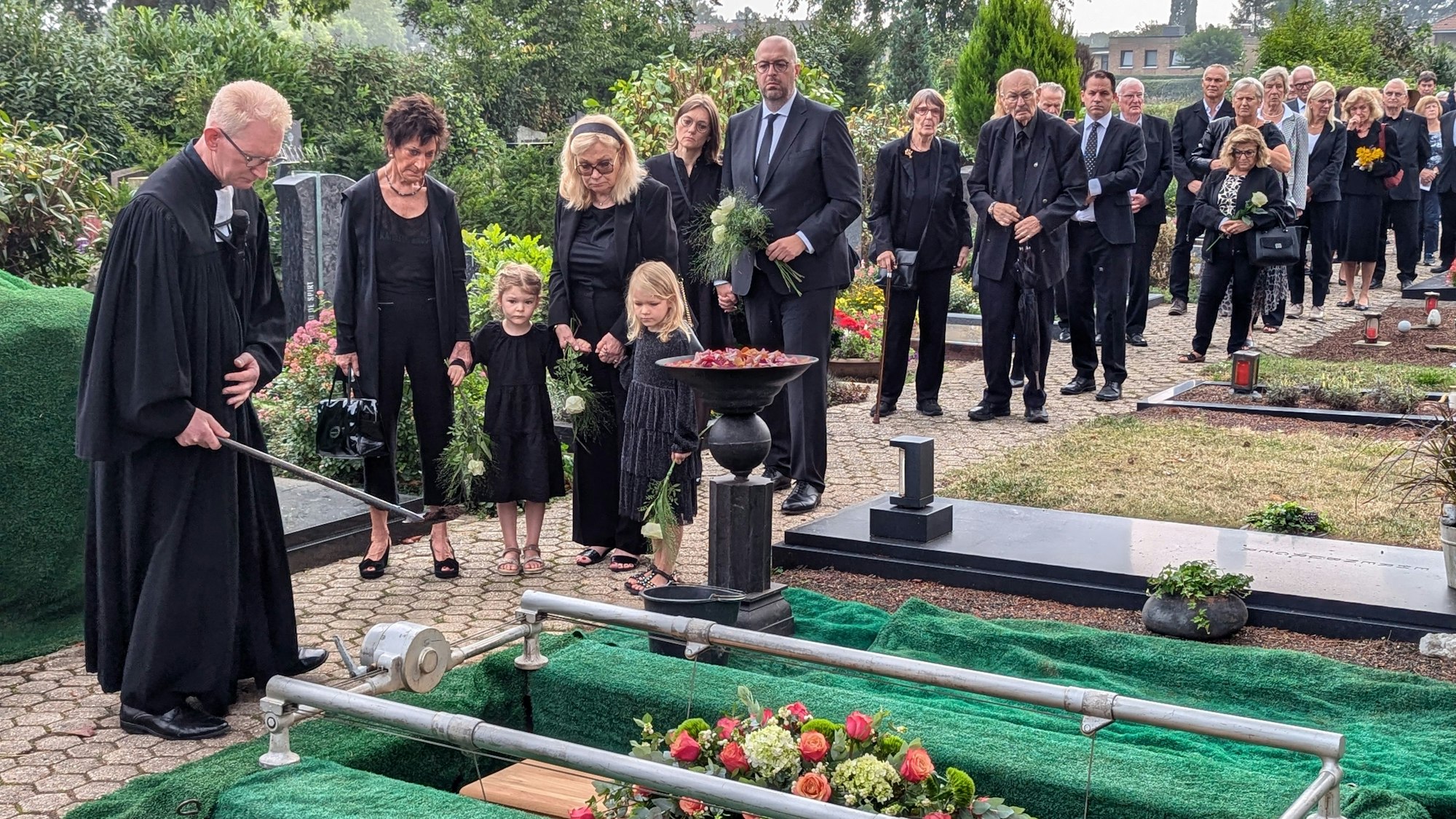 Das Foto zeigt den Pfarrer und die Familie am offenen Grab, dahinter ein langer Trauerzug.