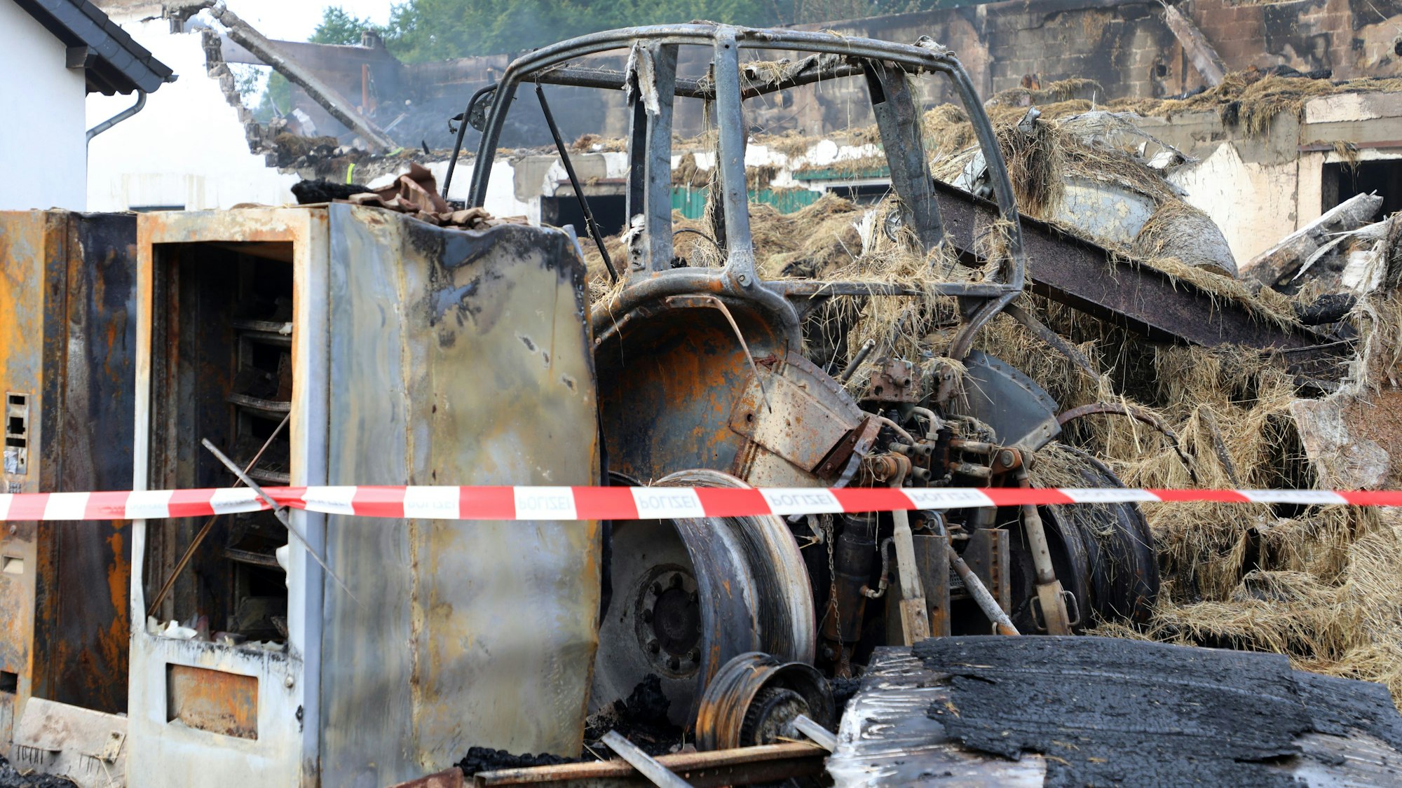 Ausgebrannte Verkaufsautomaten und ein abgebrannter Traktor stehen in Trümmern eines ehemaligen Wirtschaftsgebäudes eines Bauernhofs bei Kürten-Dürscheid.