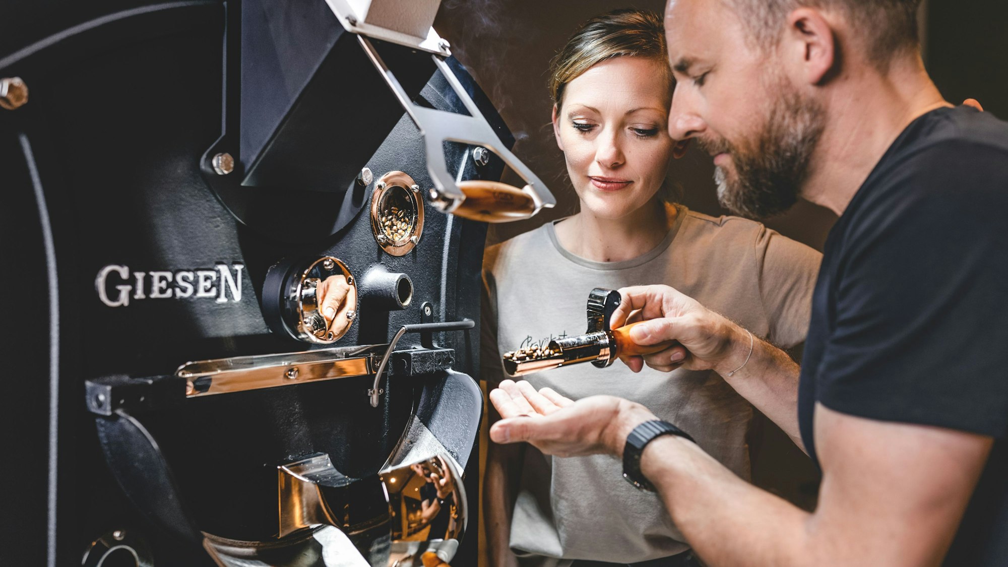 Ein Mann und eine Frau stehen vor einer Kaffeeröstmaschine mit der Aufschrift „Giesen“. In der Hand hält der Mann eine Probe mit Kaffeebohnen.