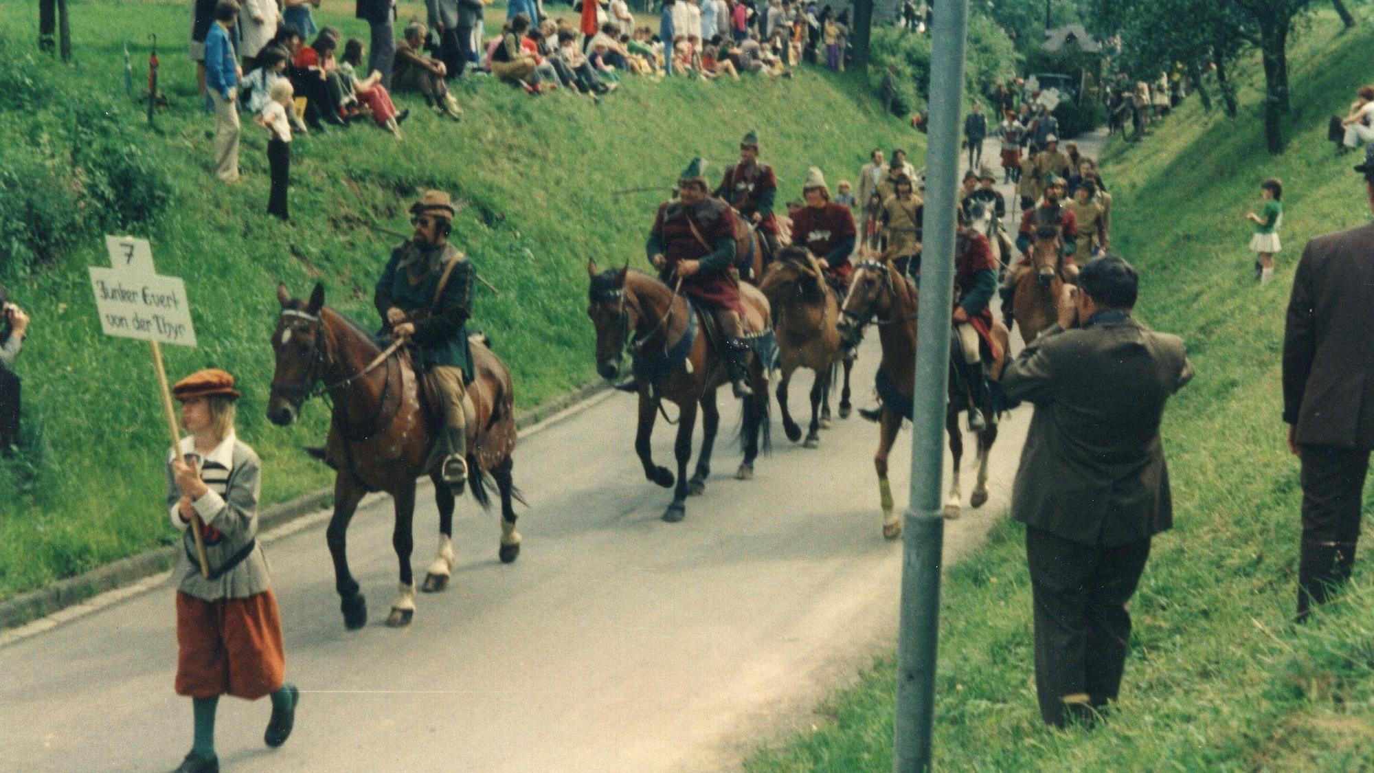 Männer in historischen Kostümen reiten auf einer schmalen Straße.
