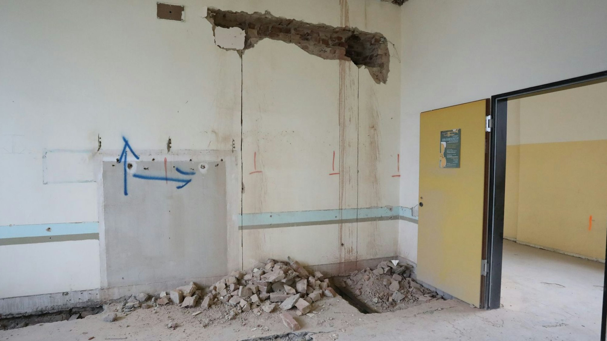 Ein leerer Raum mit geöffneter Tür, Rissen in den Wänden und Bauschutt auf dem Boden.