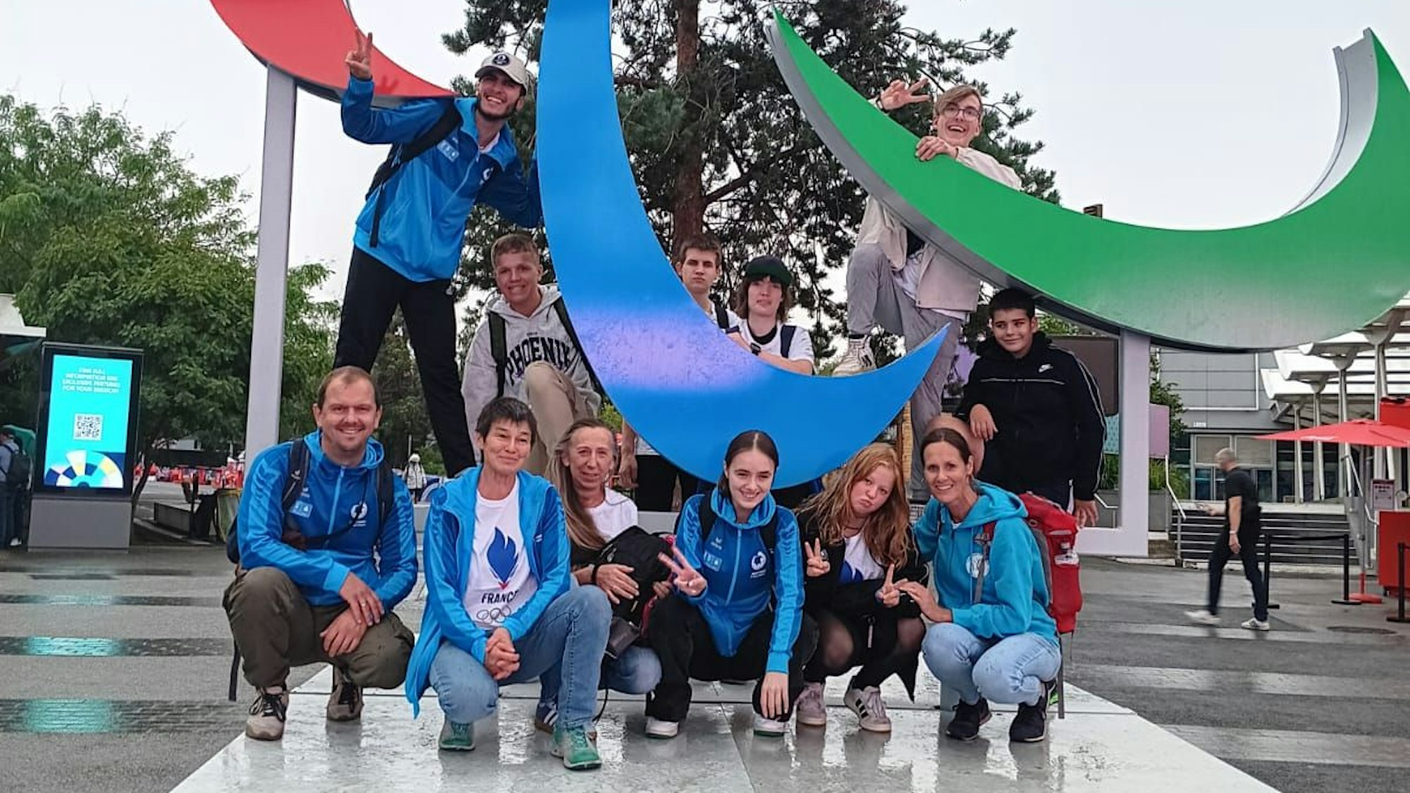 Eine Schülergruppe und ihre Betreuer stellen sich zu einem Gruppenfoto an einer Statue der Paralympischen Spiele mit der Aufschrift „Paris 2024“ zusammen.