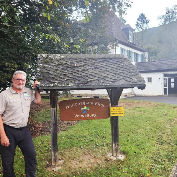 Ein Mann mit Brille steht neben einem Eingangsschild, auf dem „Nationalpark Eifel Verwaltung“ steht.