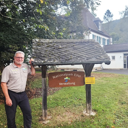 Ein Mann mit Brille steht neben einem Eingangsschild, auf dem „Nationalpark Eifel Verwaltung“ steht.