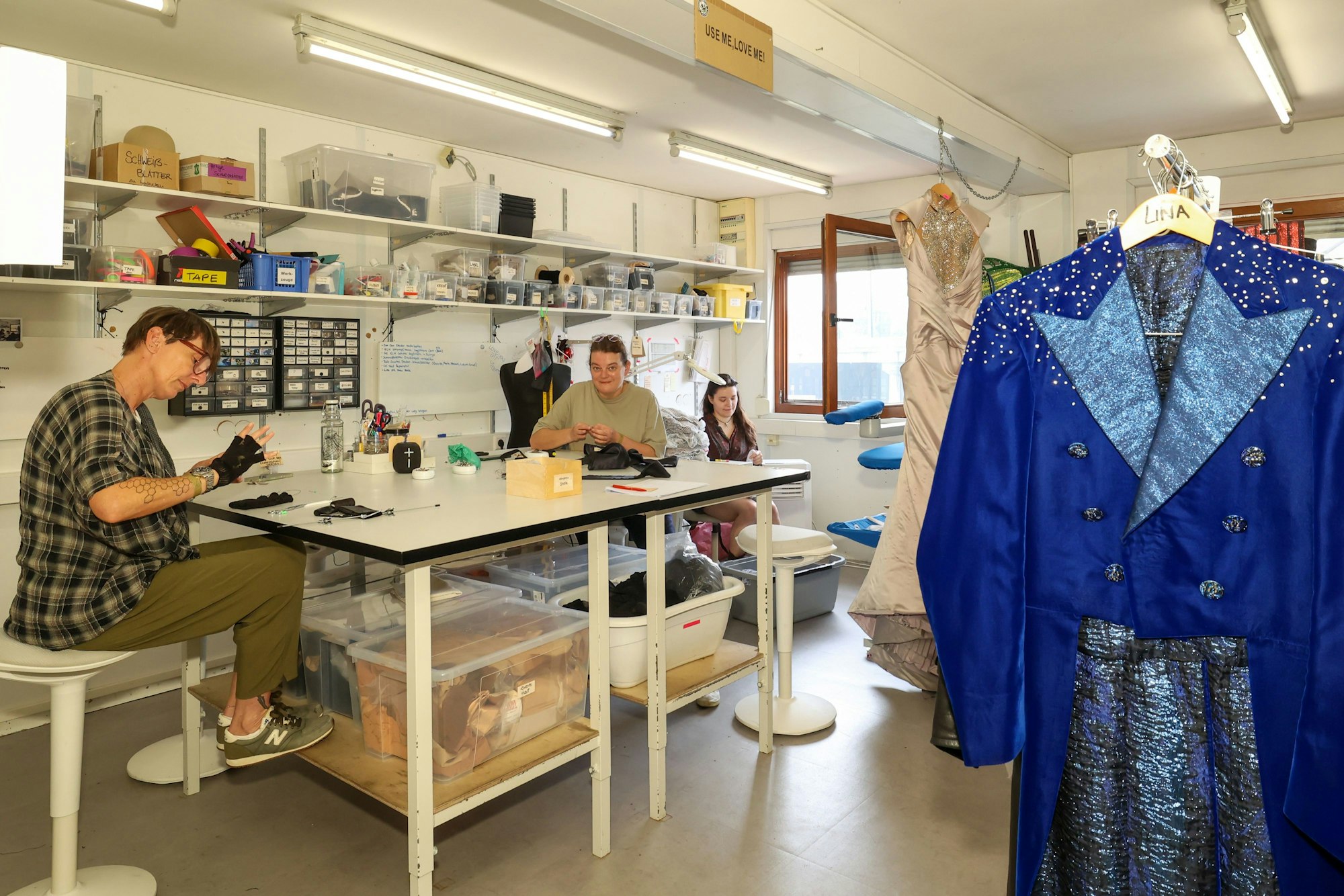 Frauen arbeiten an Kostümen, an Kleiderstangen hängen bunte und glitzernde Kostüme.
