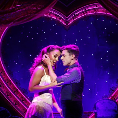 Musicaldarsteller von Moulin Rouge halten sich aneinander fest und singen, im Hintergrund ein Herz und viele kleine Lichter.