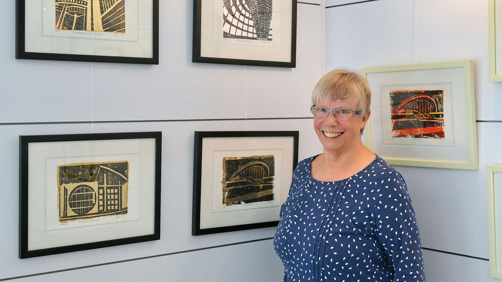Eine Frau mit Brille steht breit grinsend vor eingerahmten Bildern, die an einer Wand hängen.