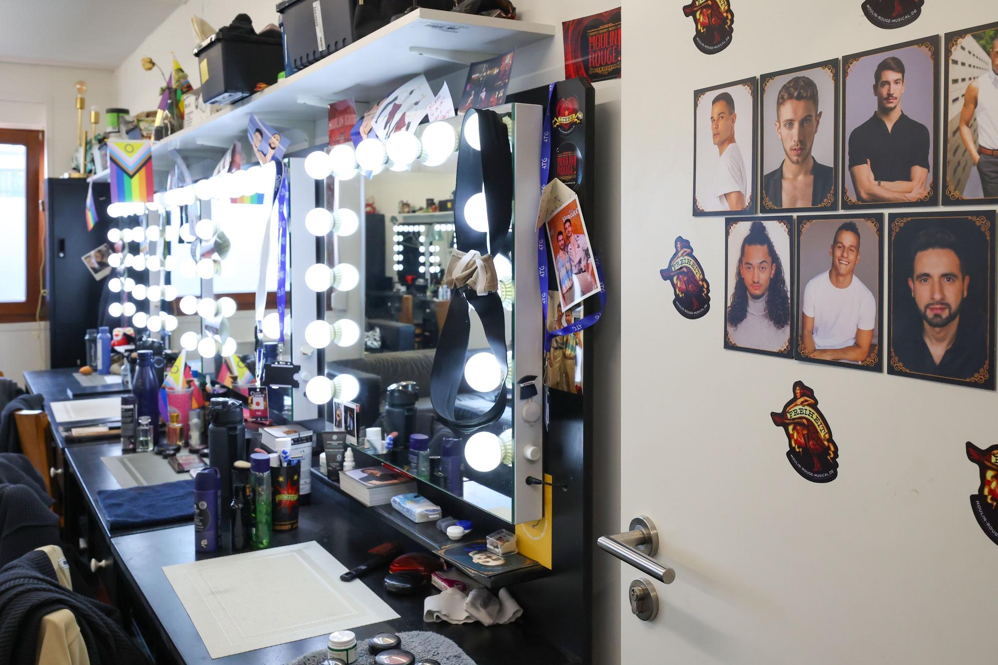 Tische stehen an der Wand, darauf Make-up- und Haarstyling-Utensilien. An der Wand hängen Spiegel mit großen Glühbirnen.