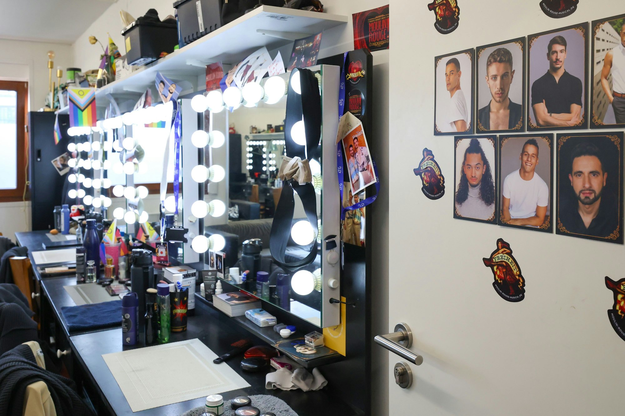 Tische stehen an der Wand, darauf Make-up- und Haarstyling-Utensilien. An der Wand hängen Spiegel mit großen Glühbirnen.