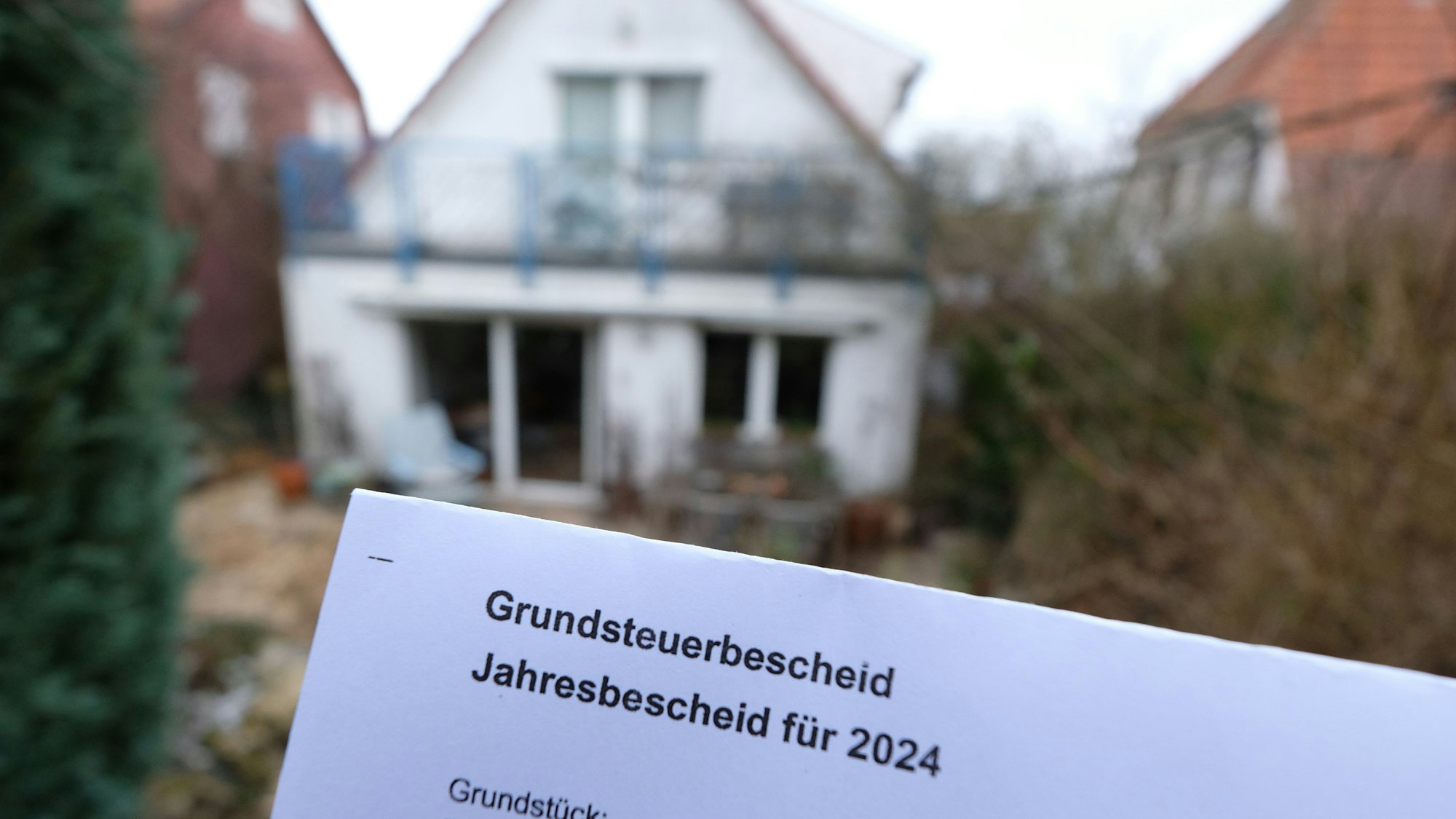 Ein Grundsteuerbescheid für 2024 wird vor einem Privatgrundstück hochgehalten.