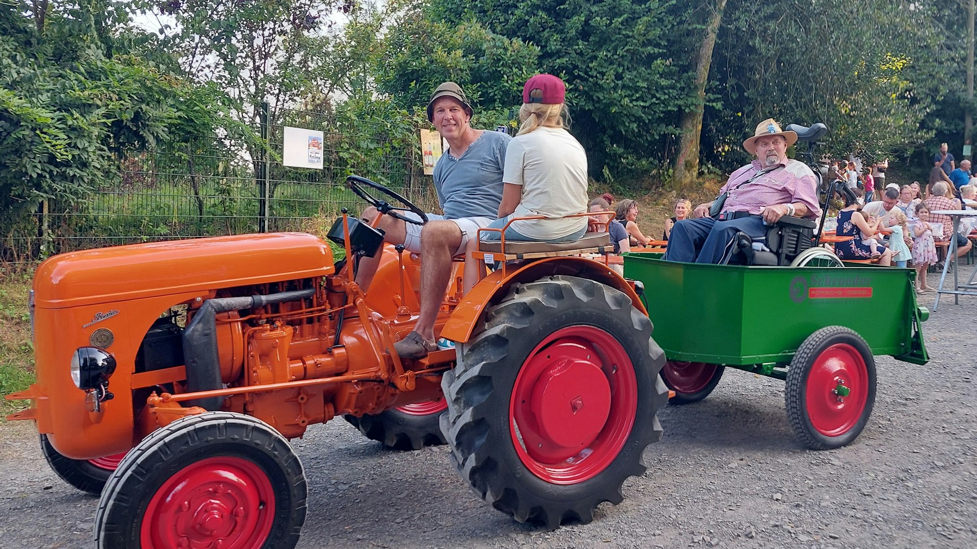 Ein orange Trecker mit Anhänger, auf dem ein Mann im Rollstuhl sitzt.