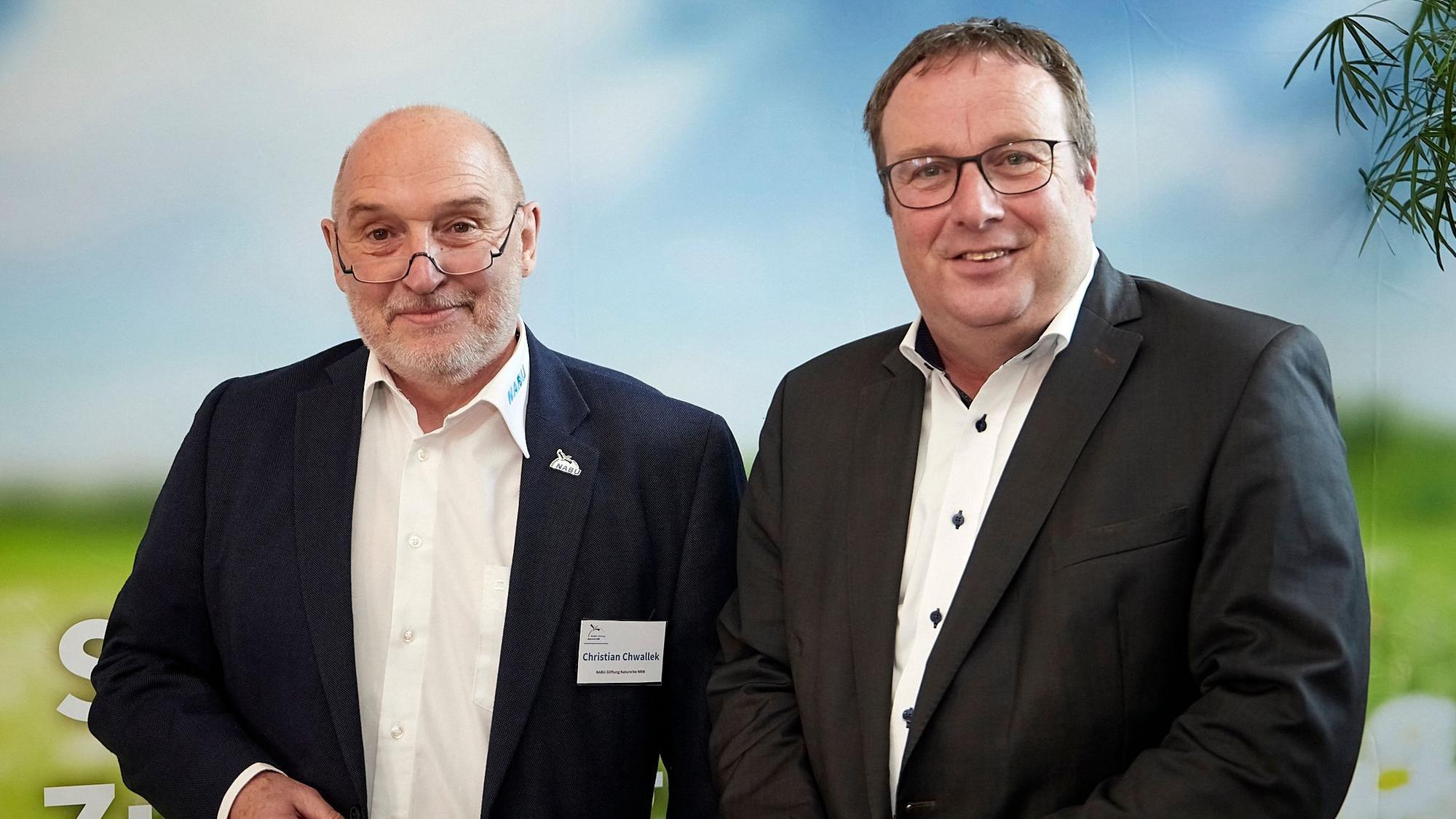 Christian Chwallek und Oliver Krischer stehen in dunklen Anzügen nebeneinander vor einem Plakat mit grünen Wiesen und blauem Himmel.