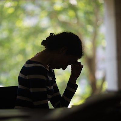 Eine Frau sitzt im Dunkeln vor einem Fenster, hat den Kopf auf einer Hand abgestützt und schaut nach unten.