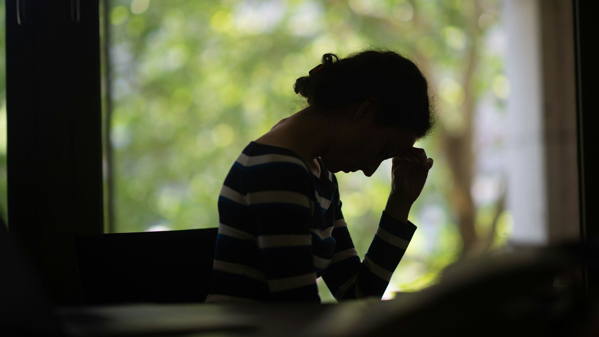 Eine Frau sitzt im Dunkeln vor einem Fenster, hat den Kopf auf einer Hand abgestützt und schaut nach unten.